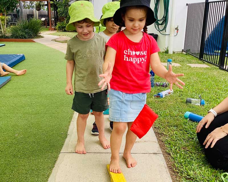 A child in a bright red shirt plays on an outdoor obstacle course while an educator sits nearby, encouraging children and verbally guiding them through challenges building their self-esteem and confidence. challenges. 