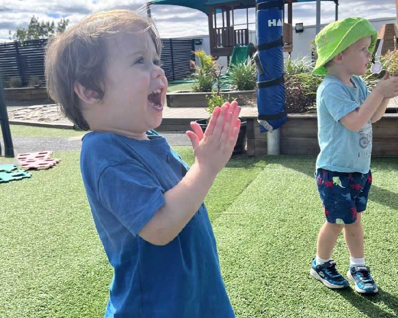Two young children enjoy the outdoors while singing nursery rhymes and moving about. Both children are clapping their hands together. Around them are wide open play spaces.