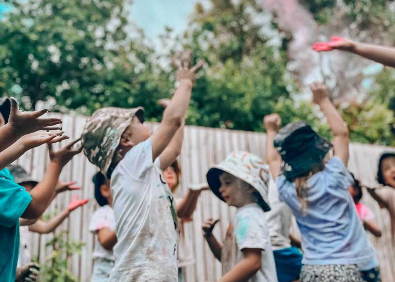 Several children gather together in an outdoor space in front of a tall fence to join in Holi the Festival of Colours' celebrations at a Petit ELJ centre. The children are all reaching up towards a cloud of pink as it rises into the air from an educator's hand seen at the top right of the photo. The children and their clothes are covered in different colours. Community celebrations help children find friends.