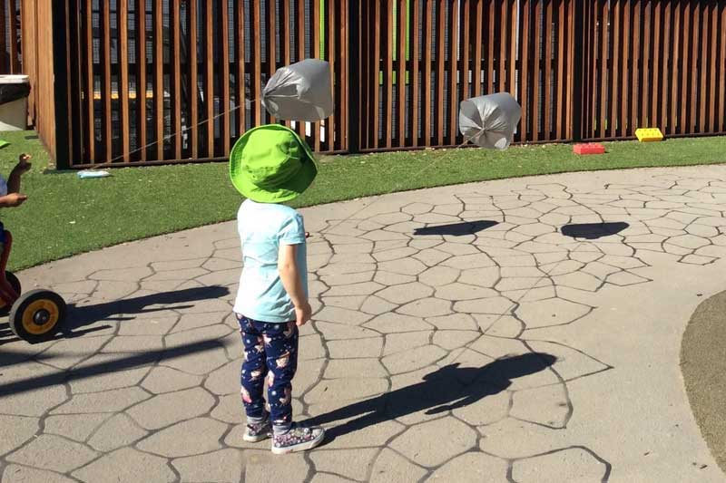 A child holds a STEM experiement of a kite made with with string and old repurposed plastic bag on a footpath in the childcare centre's outdoor environment, reaping the benefits of kite flying for children. 