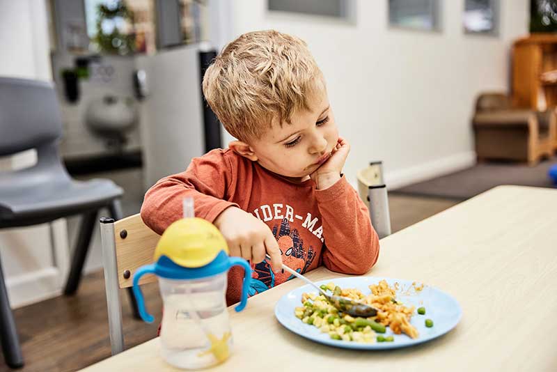 A child sits at a table eating vegetables. He looks at the plate curiously while resting a hand under his chin and using a big spoon to scoop up his meal. Next to him is his favourite yellow-topped water bottle with blue handles and a clear straw, clever choices to get children to drink water. 