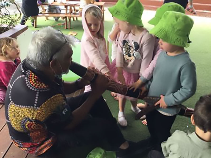 Children choose to listen and surround Indigenous elder Uncle Allan as he plays his digeridoo.