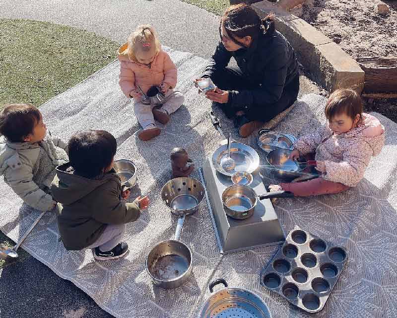 An educator sits with children on a patterned mat with repurposed man-made items for loose parts play including pats, pans, colanders, cupcake tray and other kitchen items.
