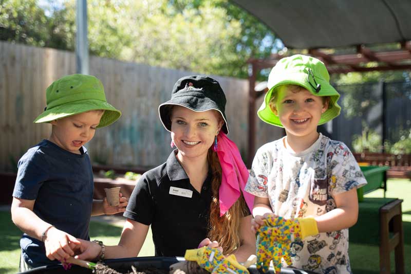 An educator growing a career with the professional development support in early childhood education and care, kneels between two children as they learn gardening together. Behind then a partially shaded outdoor setting contains more garden beds and a tall fence.