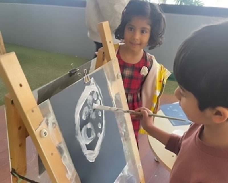 A child in a salmon coloured shirt, paints concept of planets in solar system on a black piece of cardboard with white paint on top of a wooden easel as another child in a red dress looks on for a learning project about space..
