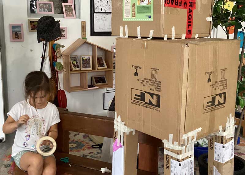 A child pulls a length of tape from a roll as she sits next to a giant cardboard creature made from several scrap boxes of different sizes and shapes, held together with masking tape. Cardboard boxes and rolls make great recycling items for preschoolers.