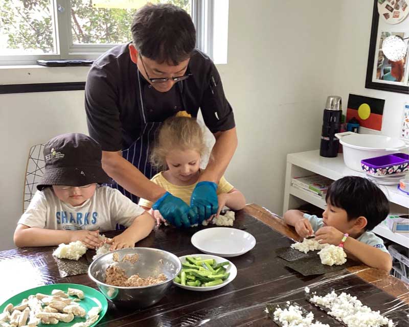  David instructs children on making sushi rolls in a cooking experience. Our chef helps a child prepare the rice while two other children squash rice on seaweed sheets. 