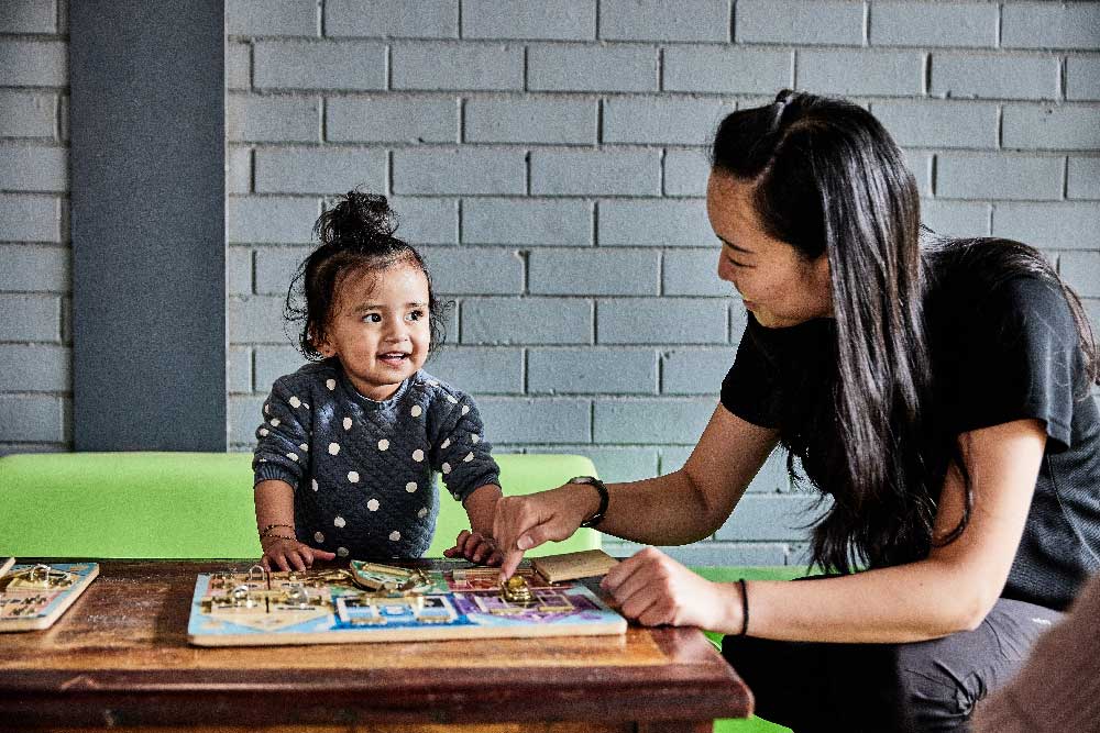 An educator sits at the child's level and points to a puzzle board game on a table. The child is standing behind the table and is smiling and looking at the educator.