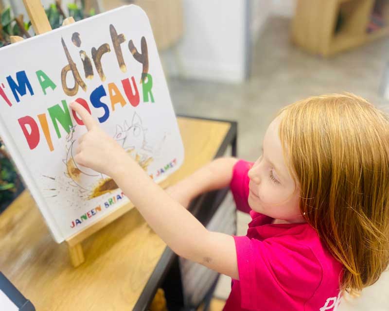 A child in a bright pink shirt points to a book on a stand with the title "I'm a dirty dinosaur". Children treasure books for bringing stories to life. 