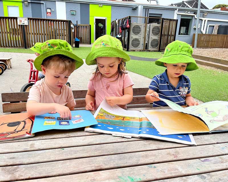 Three children wearing green Petit ELJ hats sit at an outdoor bench. Each child is looking through a large picture book reaping the benefits of storytelling. Behind them is a child on a red tricycle.
