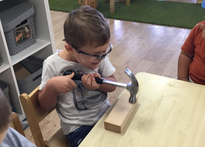 A child practises hammering on a block of wood with protection eye-wear. Involving children in making homemade keepsake boxes extends their learning experiences.