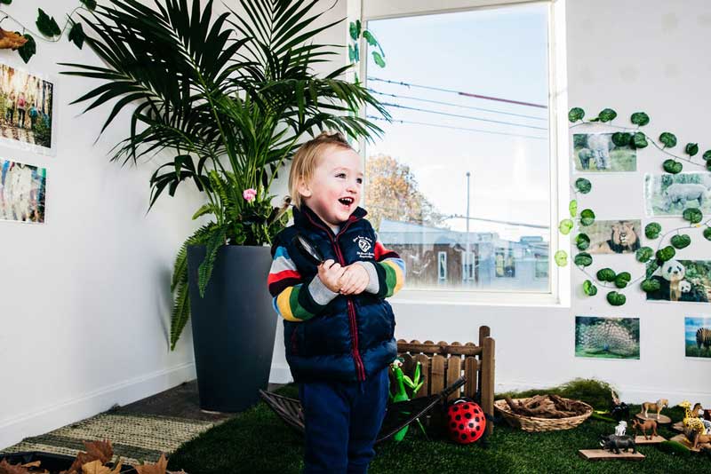 A child in a blue tracksuit stands off-centre looking towards the right of the camera while holding a magnifying glass and laughing. Behind her is a large pot plant holder with a palm and jungle-themed invitation to play.