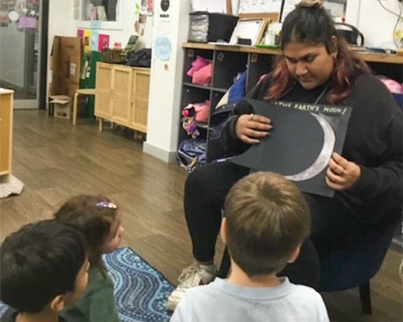 An educator sits in a chair in front of a group of kindergarten children sitting and kneeling with interest as they look at a drawing of a crescent moon. The teacher listens and responds to the children's ideas and questions about the moon as part of a space themed project learning program.