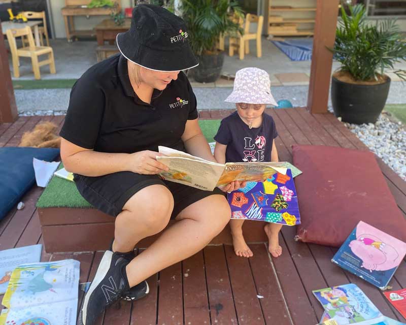 Educator Bianca from Petit ELJ Caloundra sits with a child, reading a book in out door setting surrounded by books'