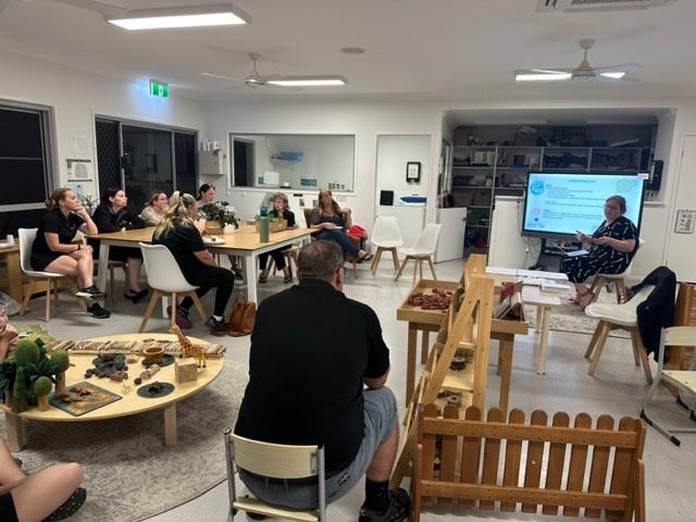 Education and Practice Advisor Emma Davis delivers training at Petit Early Learning Journey Murwillumbah to mentor and facilitate discussion on critical reflection an important aspect of the NQF. Emma sits at the whiteboard facing the studio and Murwillumbah team who gather around on desks and chair inside a studio.