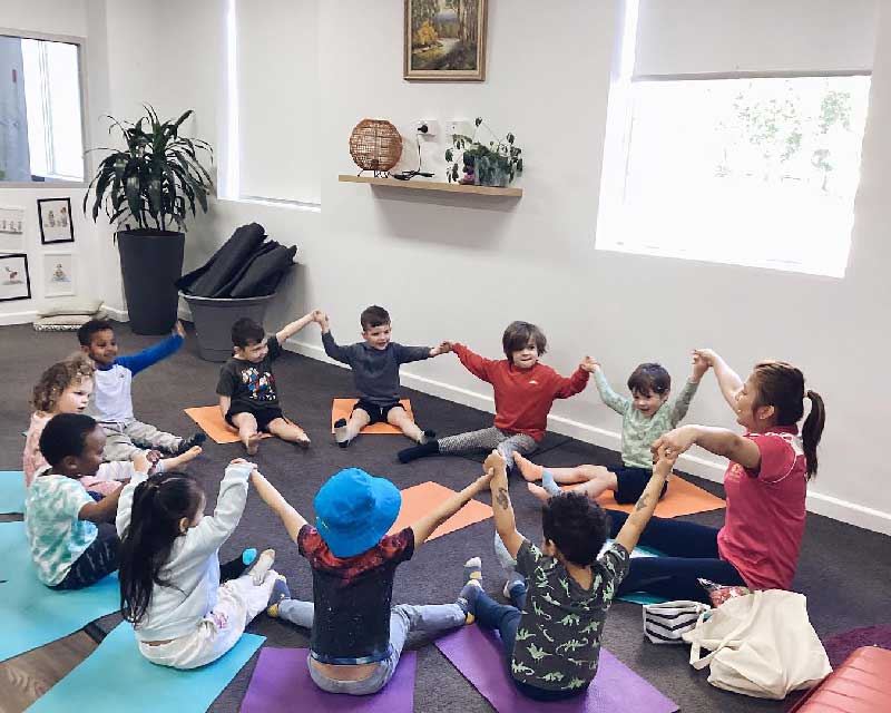 A yoga teacher and children sit inside an open studio on yoga mats. They participate in a calming activity with legs stretched out in front of them. They are connected in a circle by holding their neighbours' hands up high.