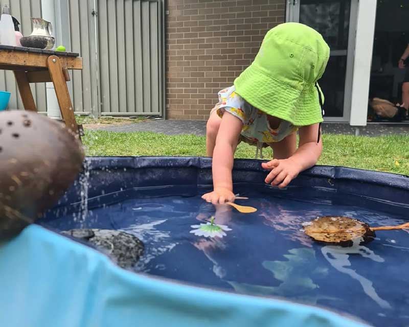 A child wearing a green hat reaches into a water-based sensory activity inside a outdoor sensory pool for a wooden spoon. There are so many sensory fun ideas for toddlers. Also in the pool is a rock, flower and wooden things. 