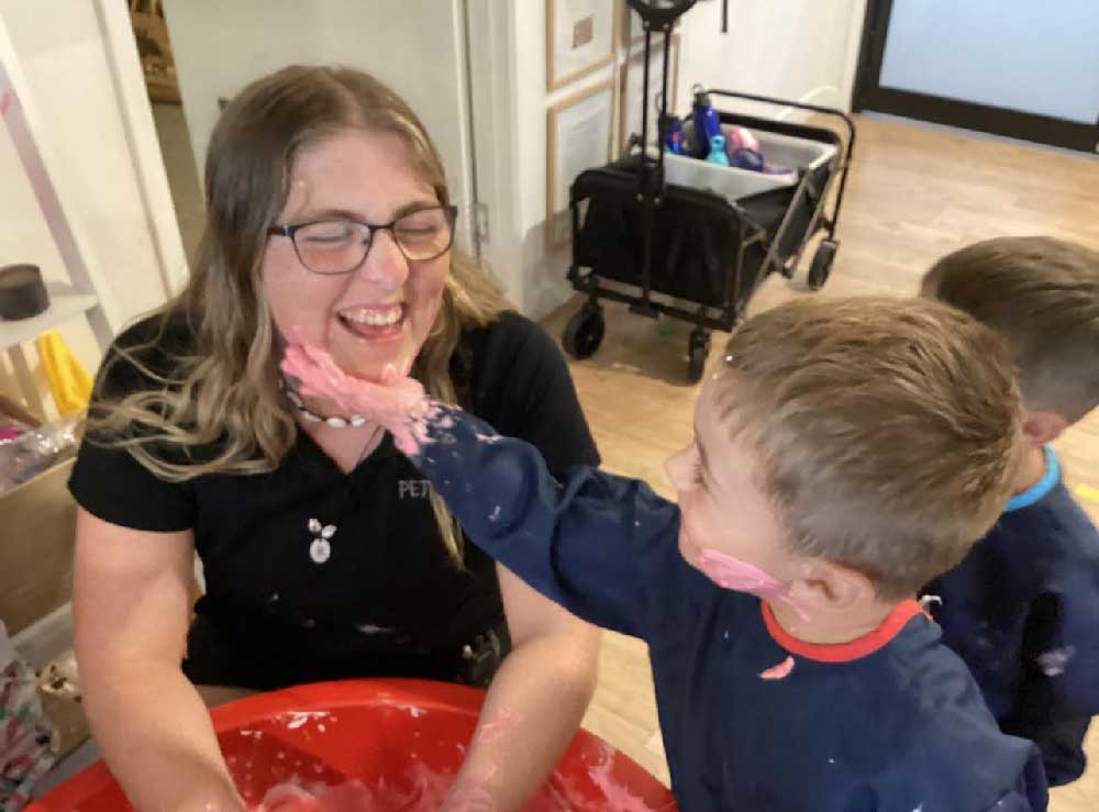 Renee McKoy, February's exceptional team member laughs and builds relationships with children at Petit ELJ Burleigh. A child with their hand covered in pink goo reaches out to place it on her face. 