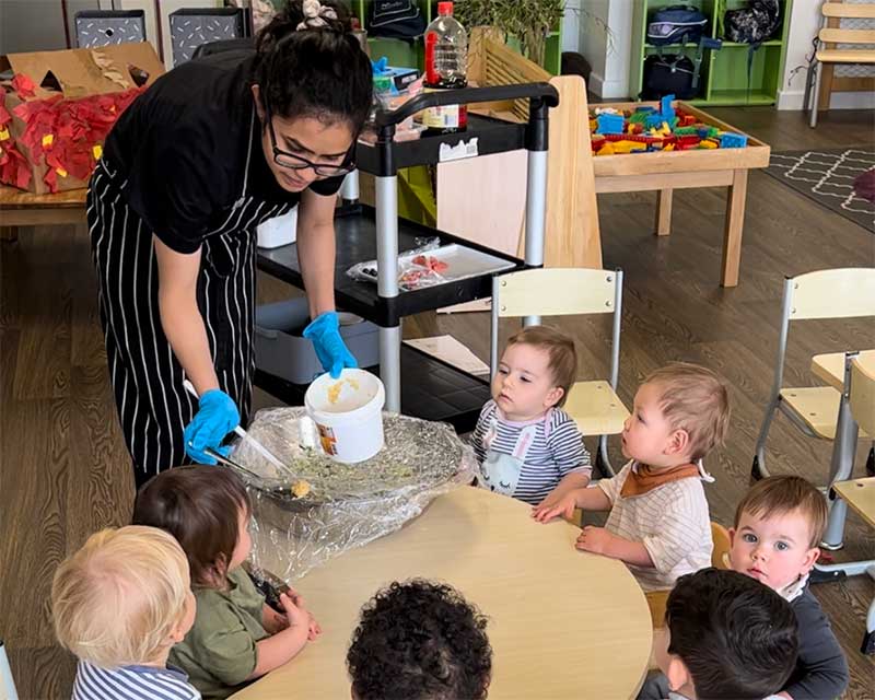 Chef Trisette who is more than just a childcare cook, hands out food for a science experiment to a group of toddlers sitting around a table. 