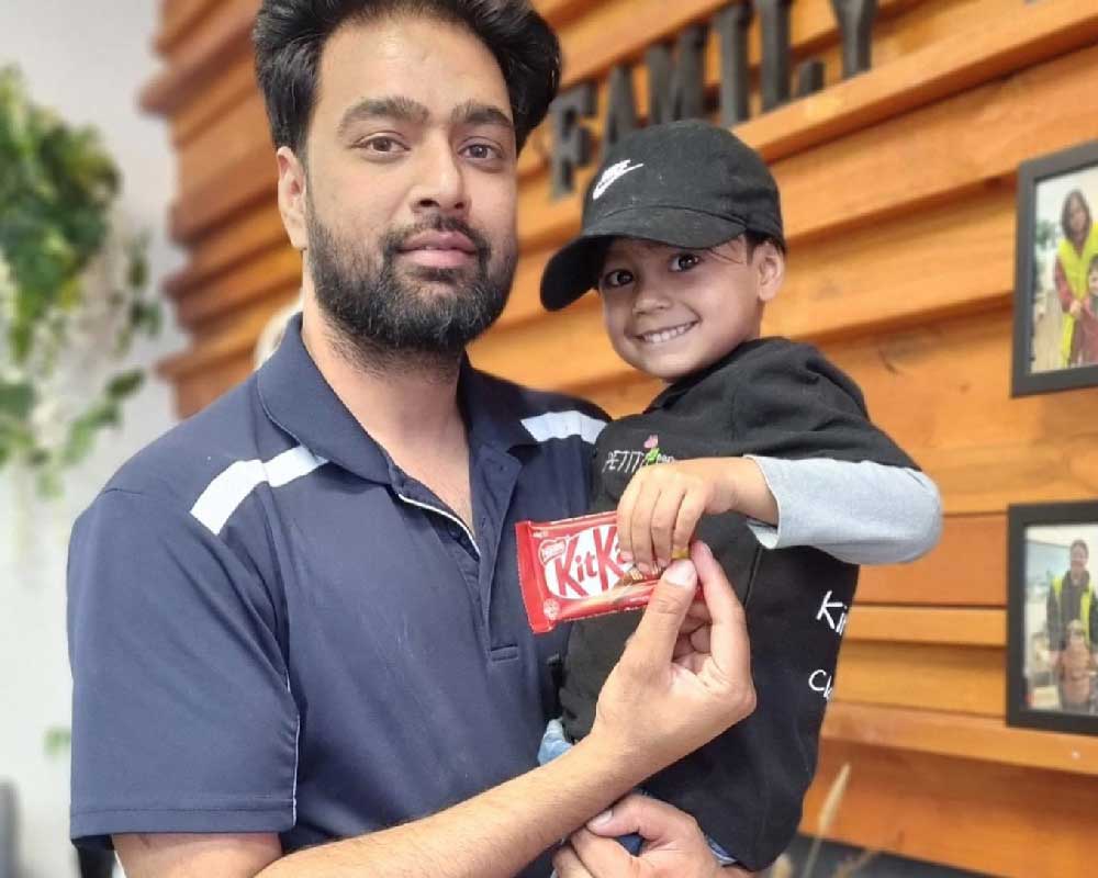 A father holds his child on Father's day at Petit Early Learning Journey. The child is and dad are holding a treat and standing in front of a wall with the word Family on it and where photos of other families are displayed.