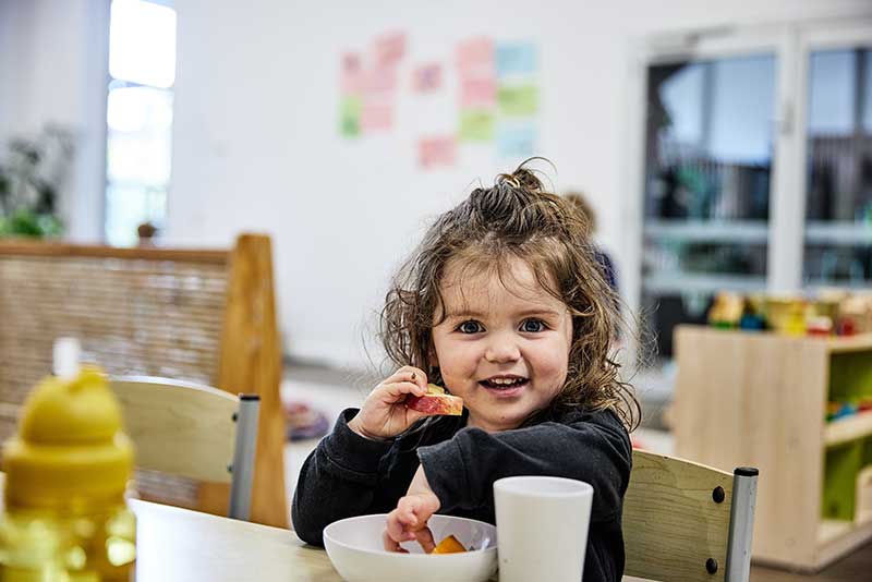 A child sits at a table eating fruit from a white bowl with a white cup beside them filled with water. In front of them is a yellow water bottle also filled with water. Children learn about the importance of water at meals, by watching adults, and exploring their world.