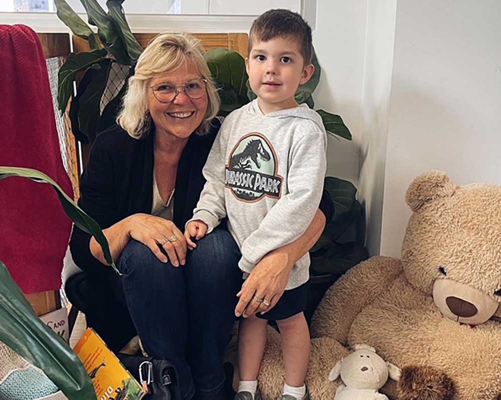 A mother and child celebrate Mother's Day just before National Families Week. The mother sits on a chair with her left arm around a child in a branded hoodie. Next to them is a giant cream coloured teddy bear.