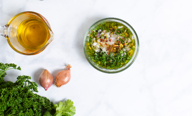 An overhead shot of a bowl of mixed fresh herbs, a cup of everyday olive oil, and a couple of shallots.