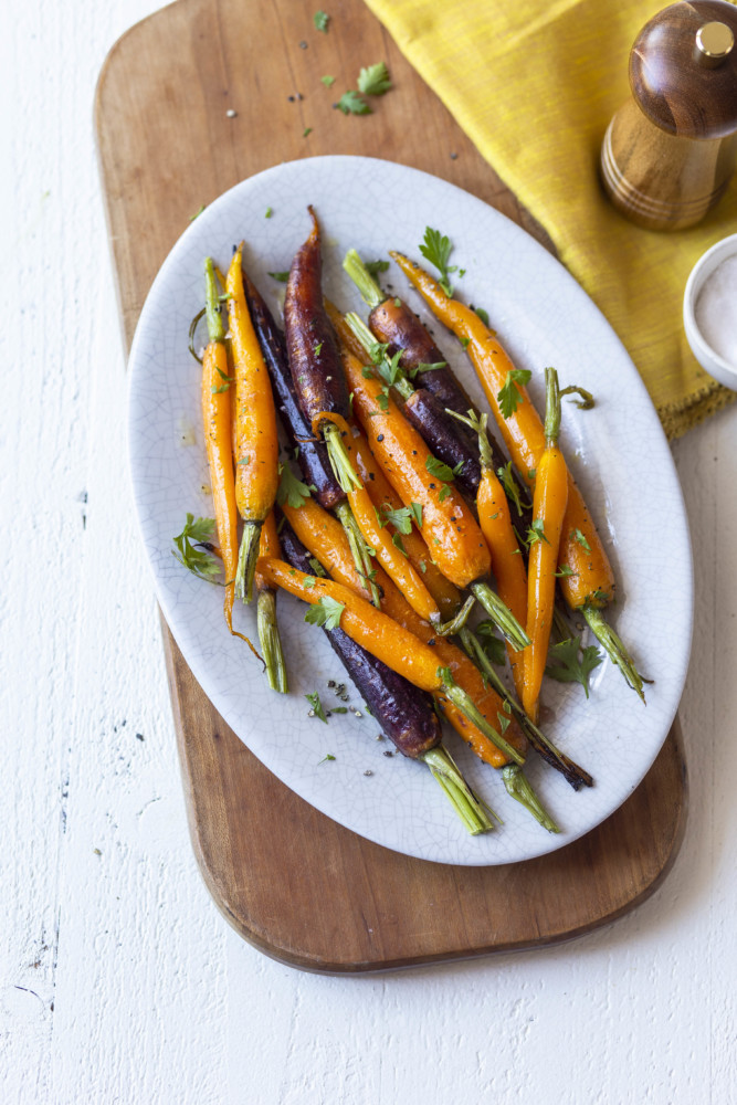 Orange and purple roasted carrots on a white plate on a wood cutting board with a pepper shaker next to it