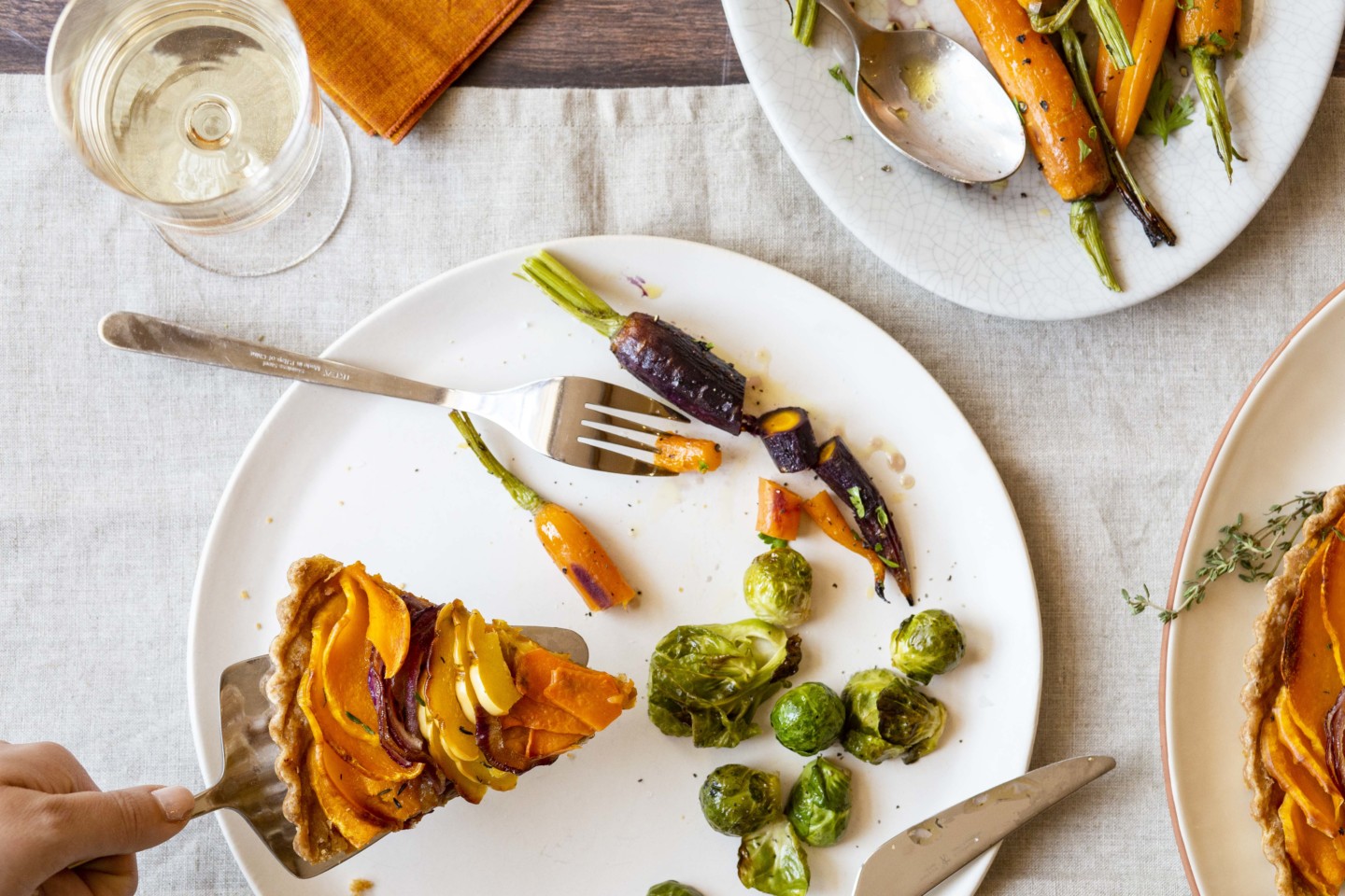 Someone serving a slice of squash tart on a plate with roasted brussels sprouts and roasted carrots. 