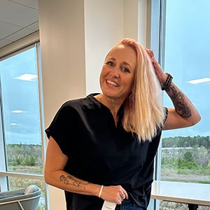 Photo of woman wearing black shirt and blonde hair smiling next to a window on a sunny day