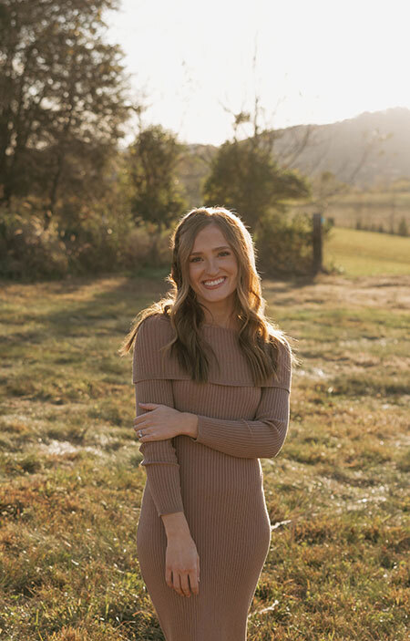 Photo of Josie Rivers who is smiling and standing outside in a grassy area wearing a brown long-sleeve dress