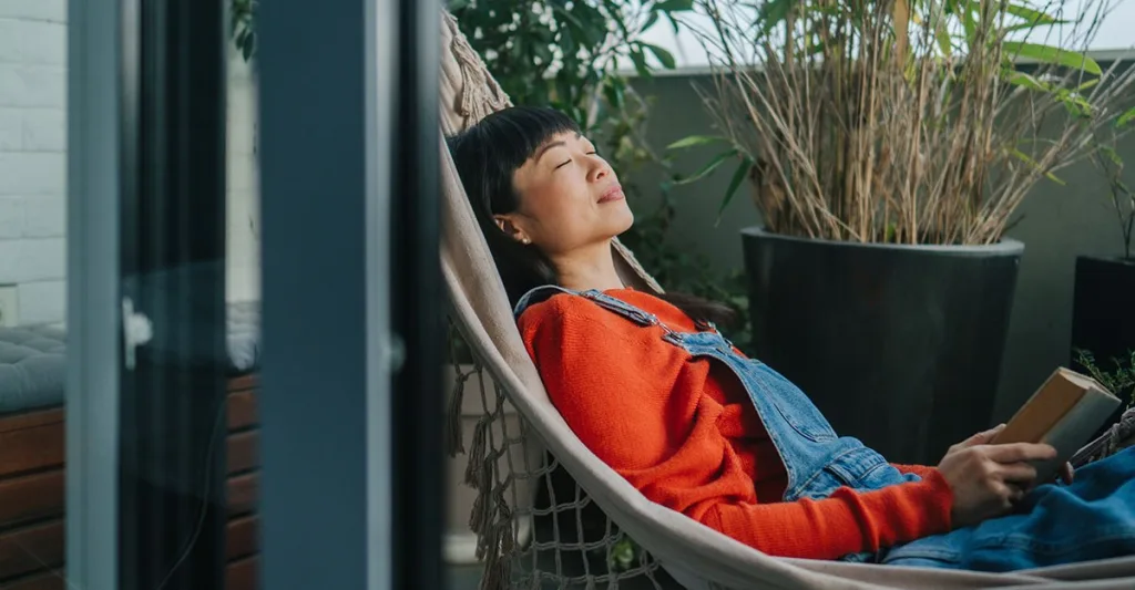 Woman practicing self-care by laying in hammock with a book with her eyes closed.