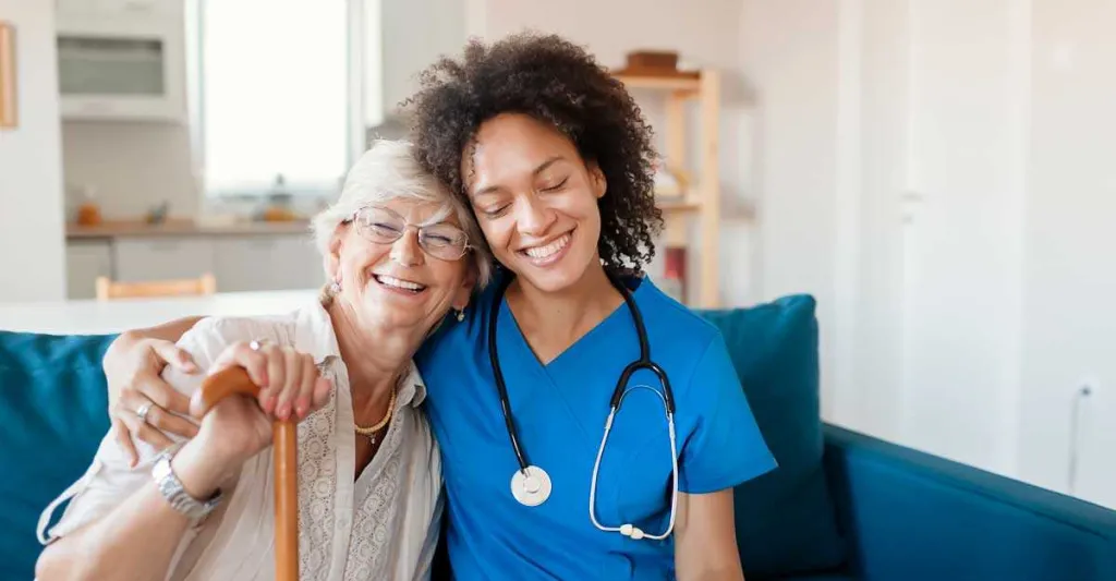 A nurse in blue scrubs sitting on a couch with an elderly patient. The nurse and patient are smiling at the camera.