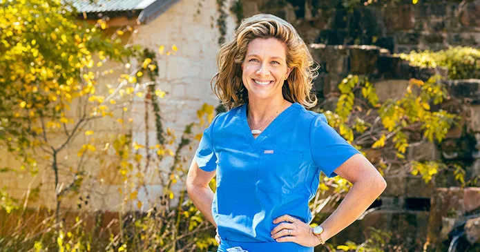 A nurse in blue scrubs smiling at the camera while standing in an outdoor setting