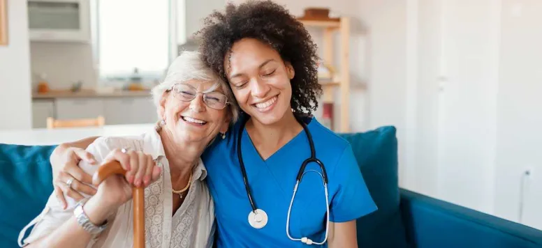 A nurse in blue scrubs sitting on a couch with an elderly patient. The nurse and patient are smiling at the camera.