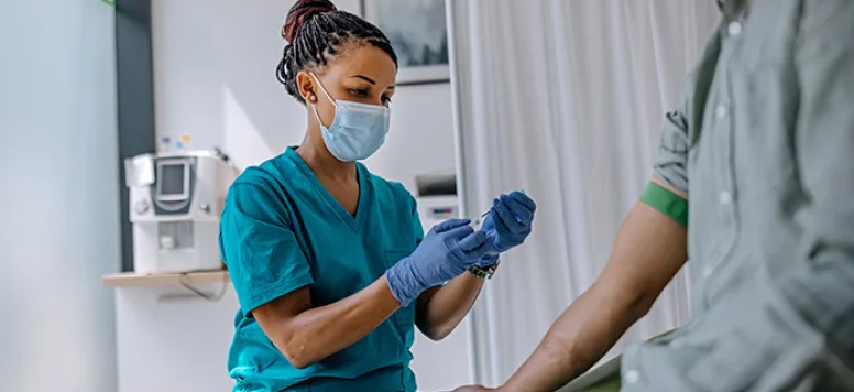 Nurse wearing surgical gloves prepares to draw blood from a patient sitting in the foreground