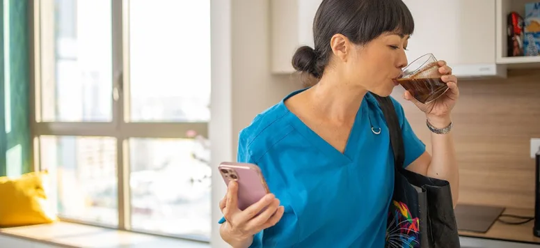 A nurse in blue scrubs carrying a handbag sips a cup of coffee in a kitchen setting