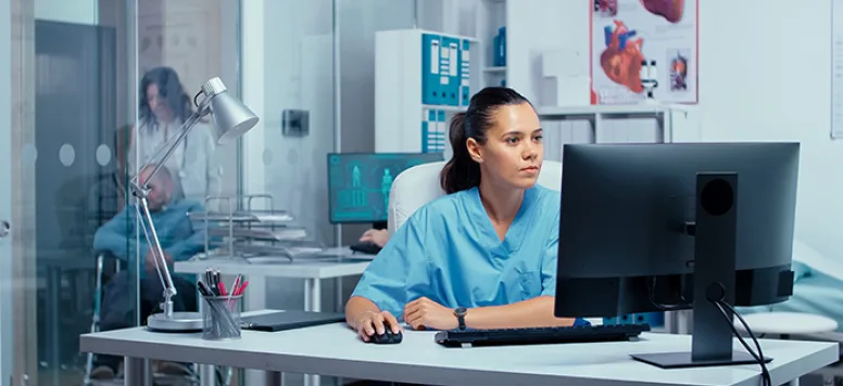 Nurse sits at desk looking at her computer while a patient sits in a wheelchair in the background