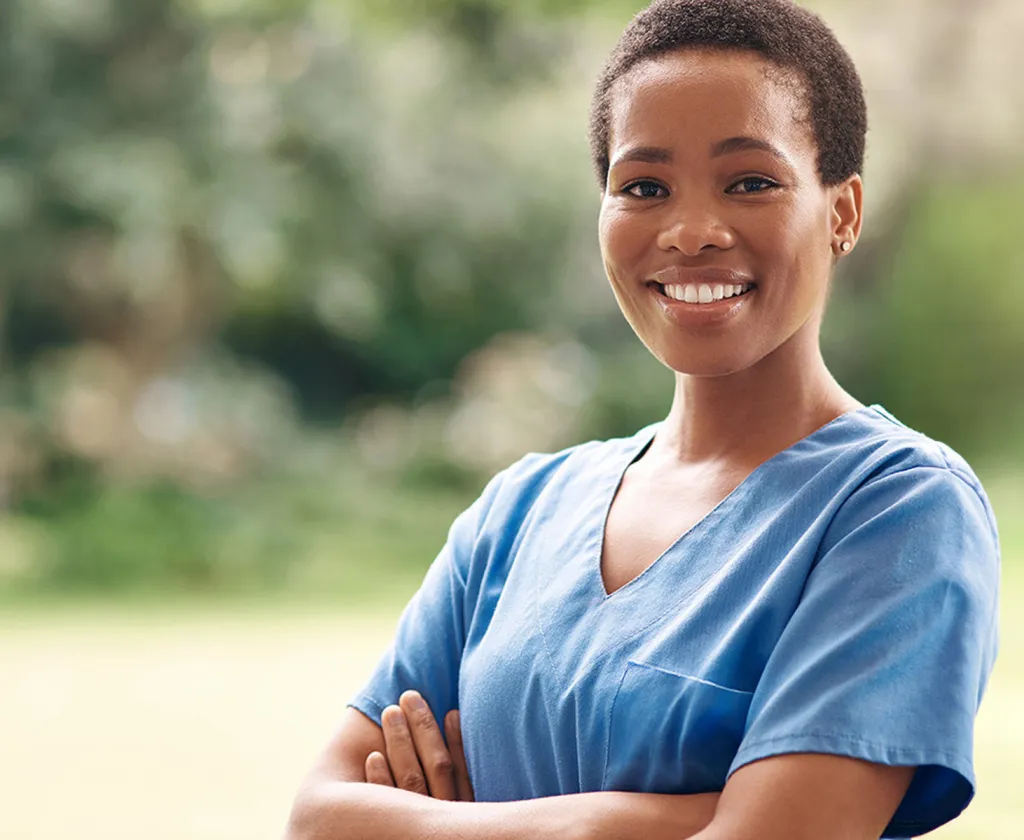 A nurse looks directly at the camera while smiling with a blurred outdoor background