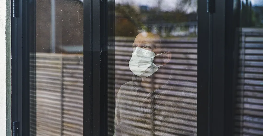 Quarantine-Man-with-surgical-mask-looking-out-window-GettyImages-1212753600.jpg