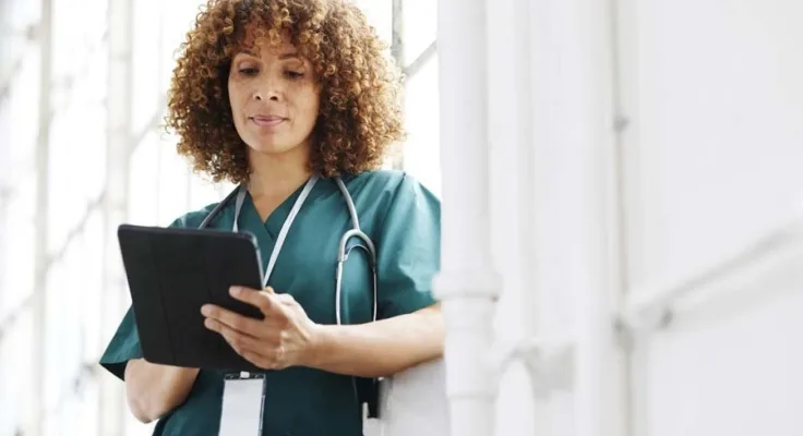 Nurse leaning on a wall reviewing a tablet