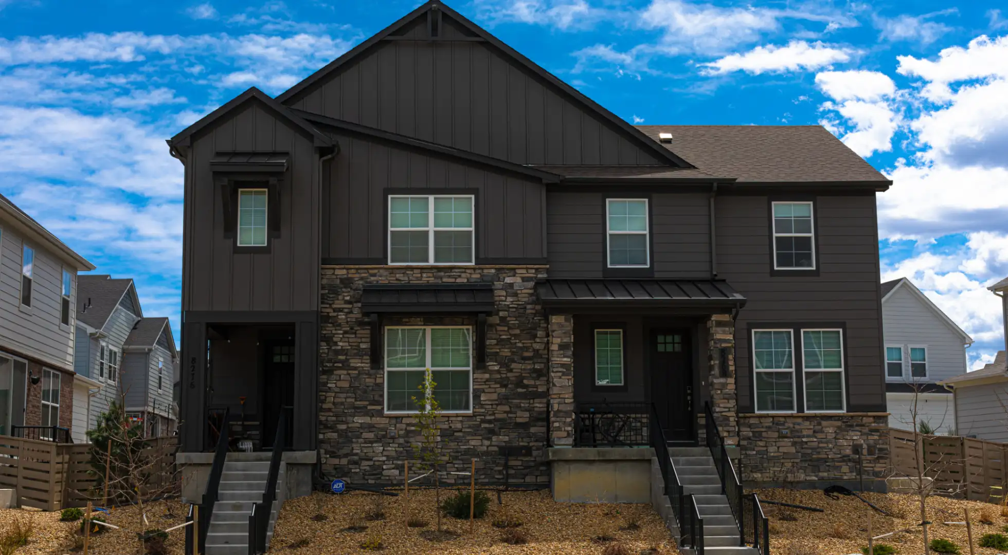 A modern two-story duplex with dark siding and stone accents under a clear blue sky.