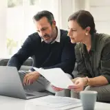 A couple discusses a document and laptop in a bright living room.