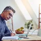 Man researching on his computer and taking notes with a pen and paper.