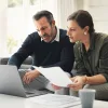 A couple discusses a document and laptop in a bright living room.