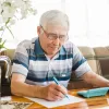 An elderly man writing at a table with a calculator nearby, using Freddie Mac's BorrowSmart program for financial planning.
