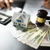 Small model of a house placed on top of a stack of U.S. dollar bills, alongside a gavel symbolizing a home foreclosure.
