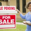 real estate agent standing next to a for sale sign with a rider reading sale pending