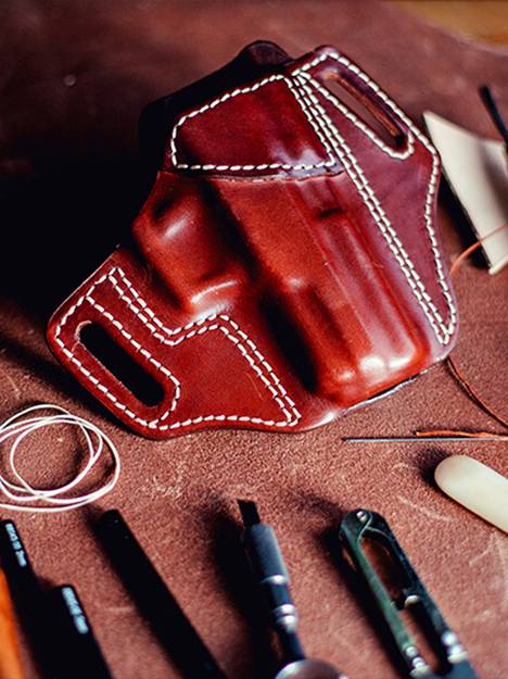 A leather OWB holster lying on a crafting desk with a lot of crafting tools around it