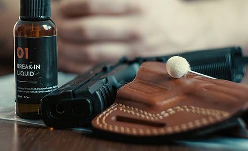 A leather OWB holster and a break-in liquid vial on a table, plastic bag underneath, a guy's arms in the background